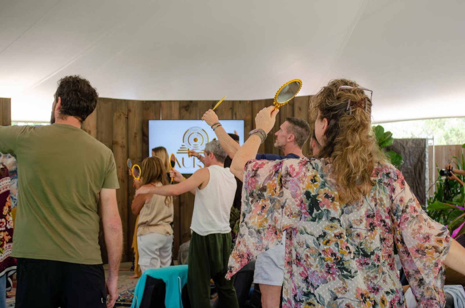 A group of people standing together under a canopy, several holding small gold hand mirrors raised.