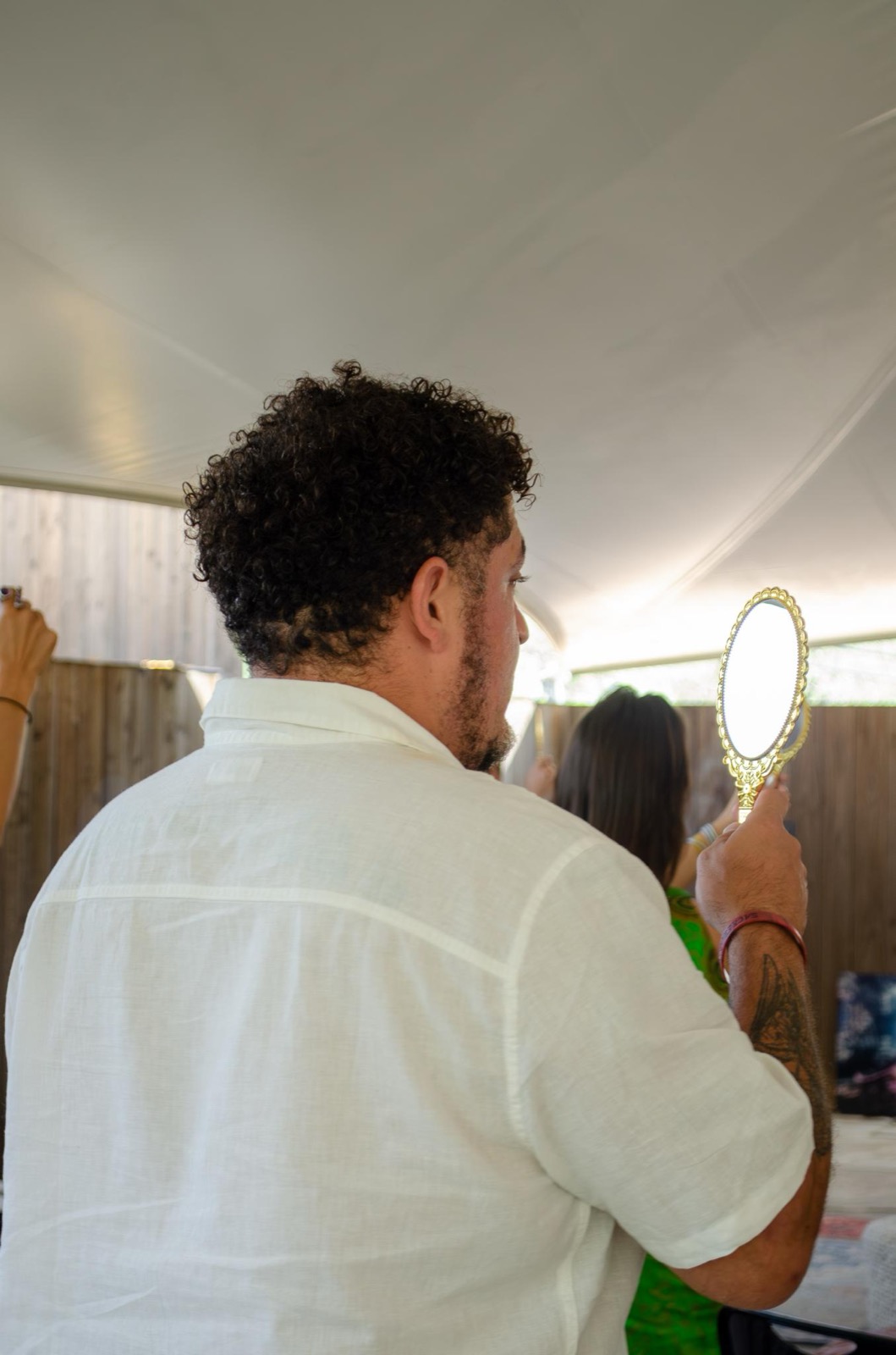 A man photographed from behind, holding a gold hand mirror up and looking into his own reflection.