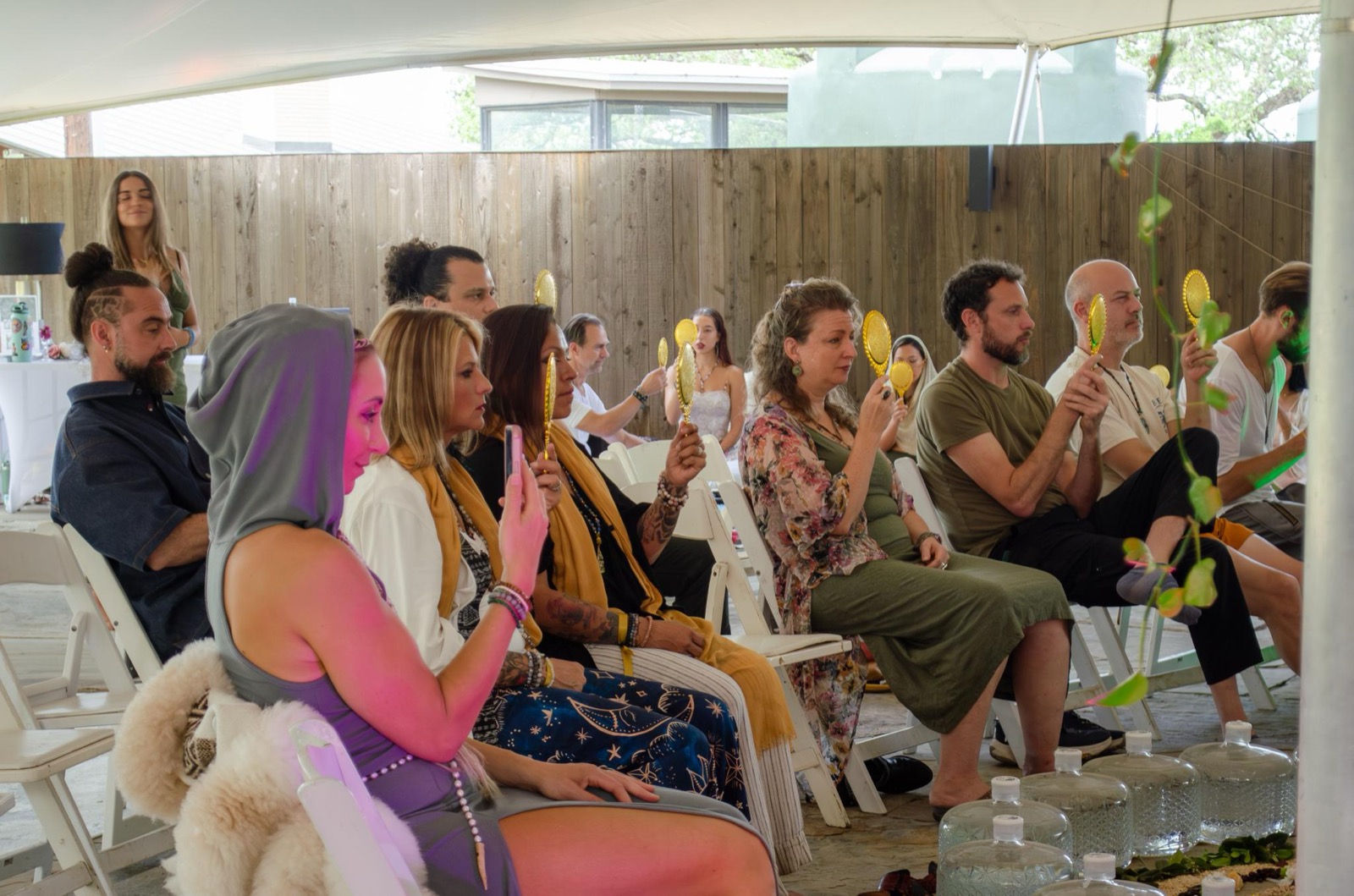 A row of people seated under a canopy, each holding a small gold hand mirror up to their own face.